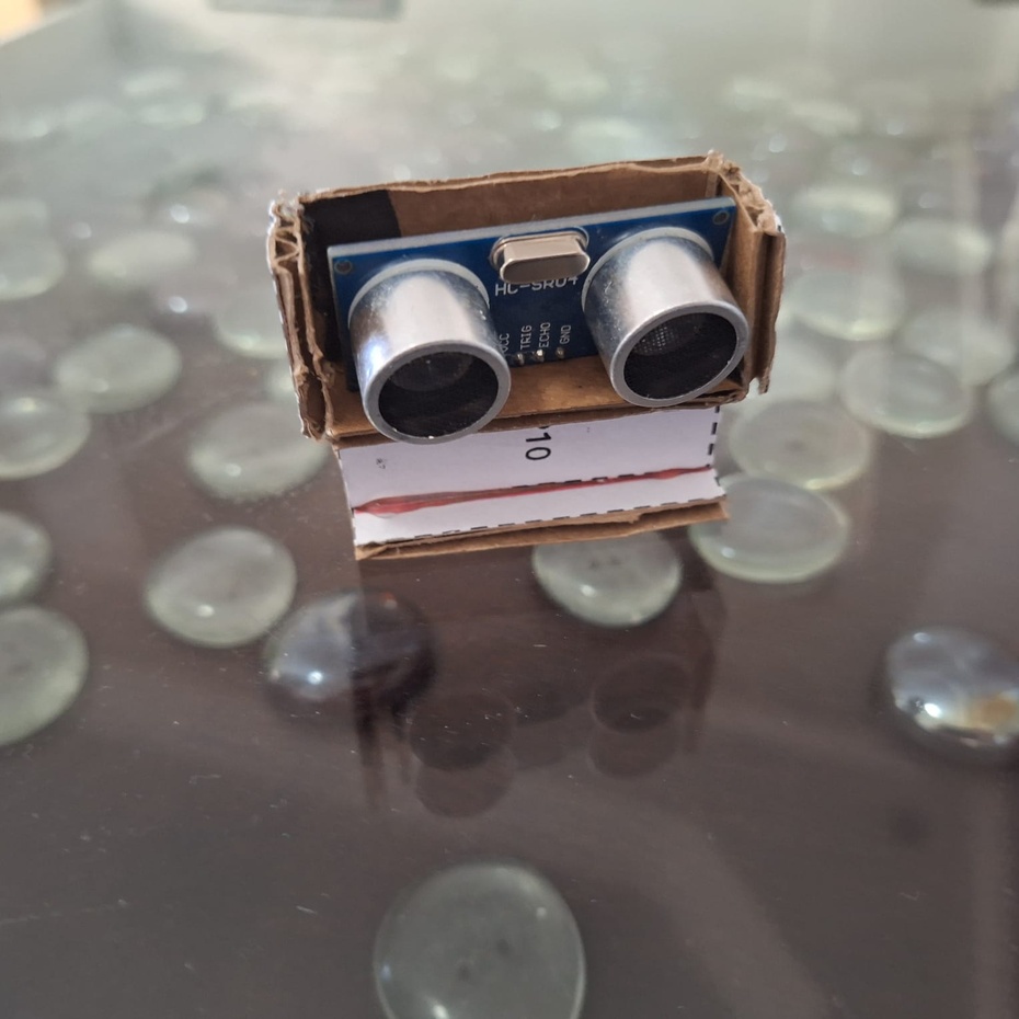 Separe as peças 6 a 11 na sua mesa de trabalho.
Cole com cola quente as peças 6, 7 e 8 com a peça 9 virada para baixo, de forma que crie uma estrutura semelhante a uma caixinha, onde o sensor de distância HC-SR04 será posicionado como na imagem abaixo.
Cole as peças 9, 10 e 11 respeitando as legendas do arquivo PPT.
No final destas colagens, seu suporte para o sensor de distância do Rolinho deve estar assim: