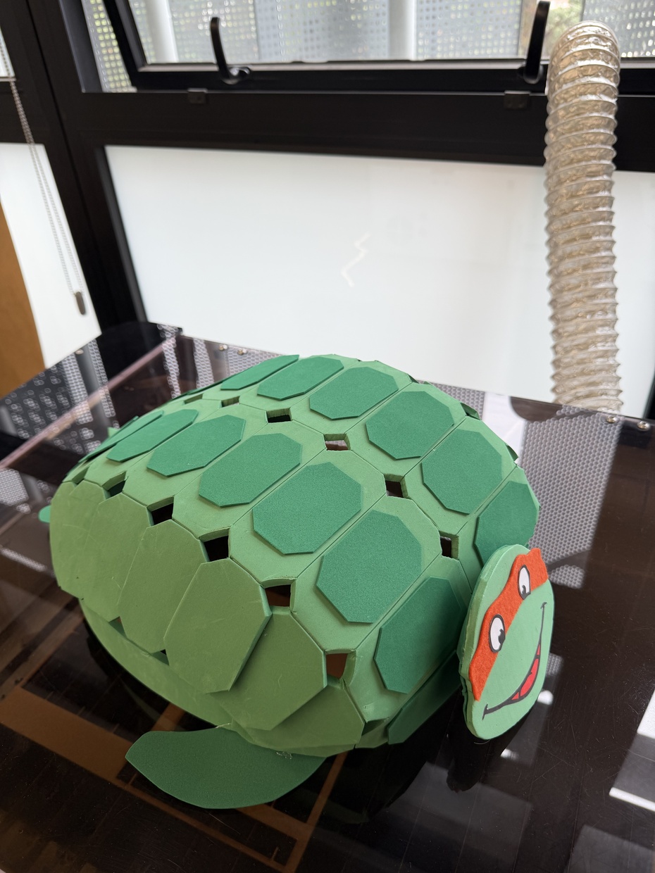 Montagem Mecânica (casco):

O casco da tartaruga foi desenvolvido artesanalmente utilizando papelão e espuma EVA, com o objetivo de criar uma estrutura leve, de baixo custo e visualmente semelhante ao casco de uma tartaruga marinha.

Inicialmente, foi construída uma estrutura curva utilizando papelão moldado manualmente e fixado sobre a base de MDF, formando o volume principal do casco. Em seguida, foram recortadas diversas peças geométricas (losangos) em EVA verde claro para compor a superfície externa, simulando as placas naturais presentes no casco de tartarugas. Sobre essas peças, foram adicionados detalhes em EVA verde escuro para criar profundidade e melhorar o acabamento visual. Todas as peças foram coladas manualmente utilizando cola quente, permitindo ajustes graduais na curvatura e alinhamento da estrutura. Durante a montagem, as peças laterais foram posicionadas em ângulo para acompanhar o formato arredondado do casco.

Além do casco principal, também foram produzidas as nadadeiras laterais e traseiras utilizando EVA recortado, fixadas na parte inferior da estrutura para reforçar a aparência de uma tartaruga marinha. A cauda foi construída com EVA em formato triangular alongado e posicionada na parte traseira do robô, complementando a identidade visual do projeto. A cabeça também foi confeccionada em EVA e personalizada com elementos inspirados no personagem Michelangelo das Tartarugas Ninja, incluindo máscara laranja, olhos e sorriso, tornando o robô mais amigável e atrativo visualmente.