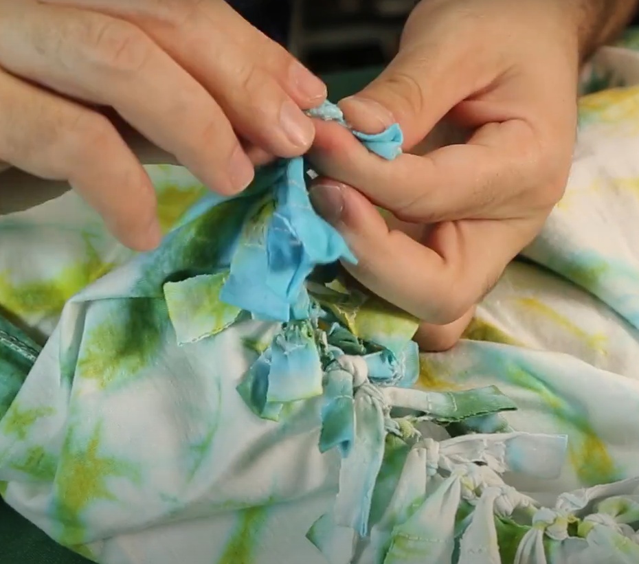 Começando pela ponta da camiseta, dê nós duplos  usando as tiras que foram cortadas. Faça isso com todas as tiras, de uma costura lateral até a outra. Lembre-se de usar as tiras correspondentes da parte da frente e da parte de trás da camiseta.