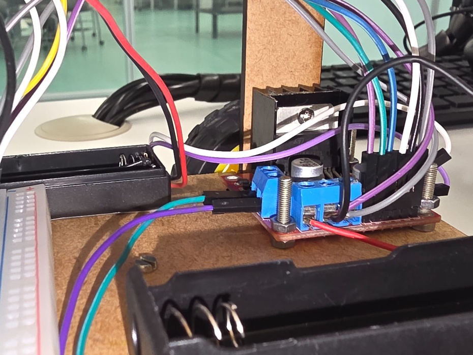 Montagem do circuito do sensor ultrassônico HC-SR04 com a adição do regulador de tensão linear LM7805

Para a montagem do circuito do sensor ultrassônico, observe que o micro:bit possui 2 portas GND. Uma delas, será utilizada para o sensor ultrassônico, enquanto a outra será ligada ao regulador de tensão linear. Dito isso, siga as seguintes especificações:

REGULADOR DE TENSÃO
- Caso esteja usando um protoboard, coloque o regulador de tensão linear de uma forma que haja ao menos uma entrada a frente e uma entrada atrás de cada pino do regulador, sendo identificados como "entrada da frente" e "entrada de trás" do respectivo pino, a partir dessa instrução;
- Pino GND do regulador de tensão linear (pela entrada da frente) é ligado à porta GND micro:bit;
- Pino GND do regulador de tensão linear (entrada de trás) é ligado à entrada GND da ponte-H. Haverá 2 fios ligados a essa porta: um fio do suporte de pilha (identificado como fio preto na imagem abaixo) e um fio jumper do regulador de tensão (identificado como fio roxo na imagem abaixo);
- Pino IN do regulador de tensão é ligado a entrada 5V da ponte-H (identificado como um fio cinza na imagem abaixo);
- Pino OUT do regulador de tensão é ligado ao pino VCC do sensor ultrassônico.

SENSOR ULTRASSÔNICO
- Pino VCC do sensor ultrassônico é ligado ao pino OUT do regulador de tensão;
- Pino GND do sensor ultrassônico é ligado à porta GND do micro:bit;
- Pino TRIG do sensor ultrassônico é ligado à porta 15 do micro:bit;
- Pino ECHO do sensor ultrassônico é ligado à porta 16 do micro:bit.

Observação: A imagem abaixo mostra dois fios ligados à entrada GND da ponte-H L298N, sendo o fio preto proveniente do suporte de pilhas e o fio roxo proveniente do pino GND do regulador de tensão linear.