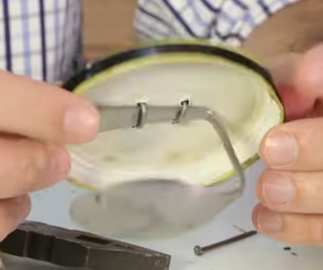 Fixe o cabo da colher (oposto à ponta) no interior da tampa, e para isso utilize os pedaços de arame dobrados, passando as pontas pelos furos feitos anteriormente (de dentro para fora da tampa), e para que fiquem fixos, abraçando a colher e a tampa, enrole as pontas de arame uma na outra, as dobre sobre a tampa e finalize colando fita isolante nessa parte externa da tampa, ajudando a fixar o arame no lugar e também fechando os buracos por onde os arames passaram.