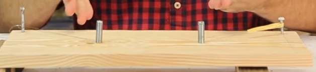 Finalize prendendo o outro lado do laço ao último parafuso que sobrou (próximo a borda da madeira), para fazer isso, passe um elástico por dentro do laço, o dobre ao meio e então passe o parafuso por dentro das duas metades do elástico. O importante é que a corda de violão fique bem esticada e firme, se um elástico não for suficiente, utilize mais elásticos, da mesma forma, até que sinta que a corda ficou firme.